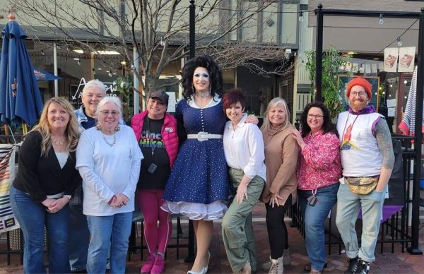 Nine people pose for a photo outside, including a drag queen wearing a blue dress in the center.